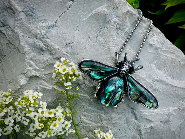 A turquoise beetle shaped crystal necklace ontop of a stone nest to some white flowers